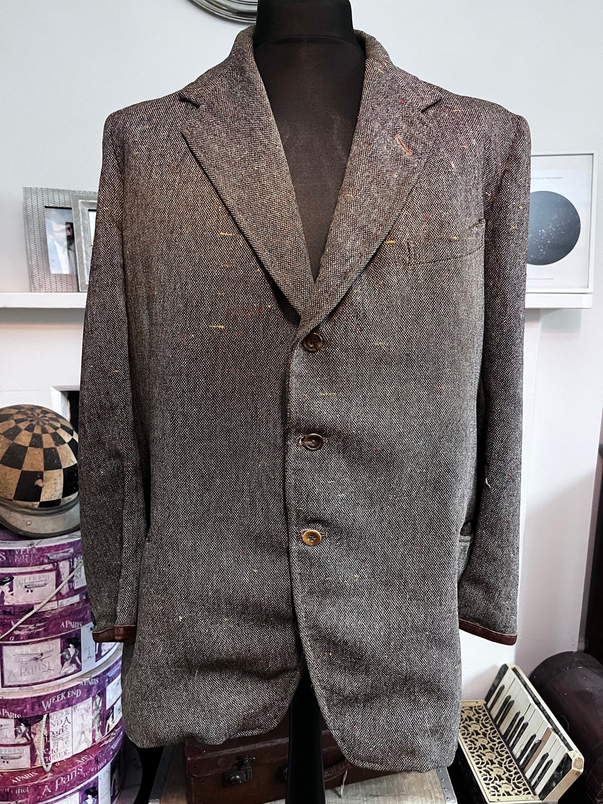 May include: A brown tweed blazer with a single-breasted closure and three buttons. The blazer has a notched lapel and brown leather trim on the sleeves.